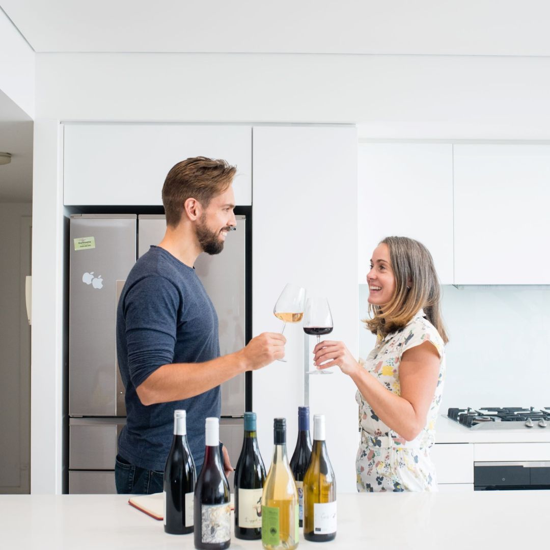 A photo of founder Mike and his partner Melissa holding wine glasses with six bottles of Feravina wines on a kitchen bench in front of them
